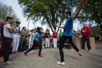 Two players circle around each other at a capoeira event at Stanford University, while onlookers sing and clap.