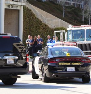 A group of fire officials is gathered in front of police and fire vehicles. One has his head down. Another holds a bottled beverage.