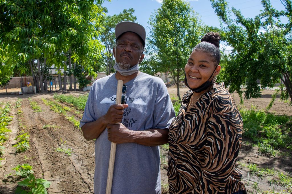 A man and woman are looking at the camera, they have their masks on their chin. They're standing in a yard.