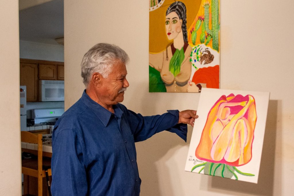 A man with gray hair hold sup a painting of a flower. In the background is another painting of a woman with the Mexican flag and cacti.