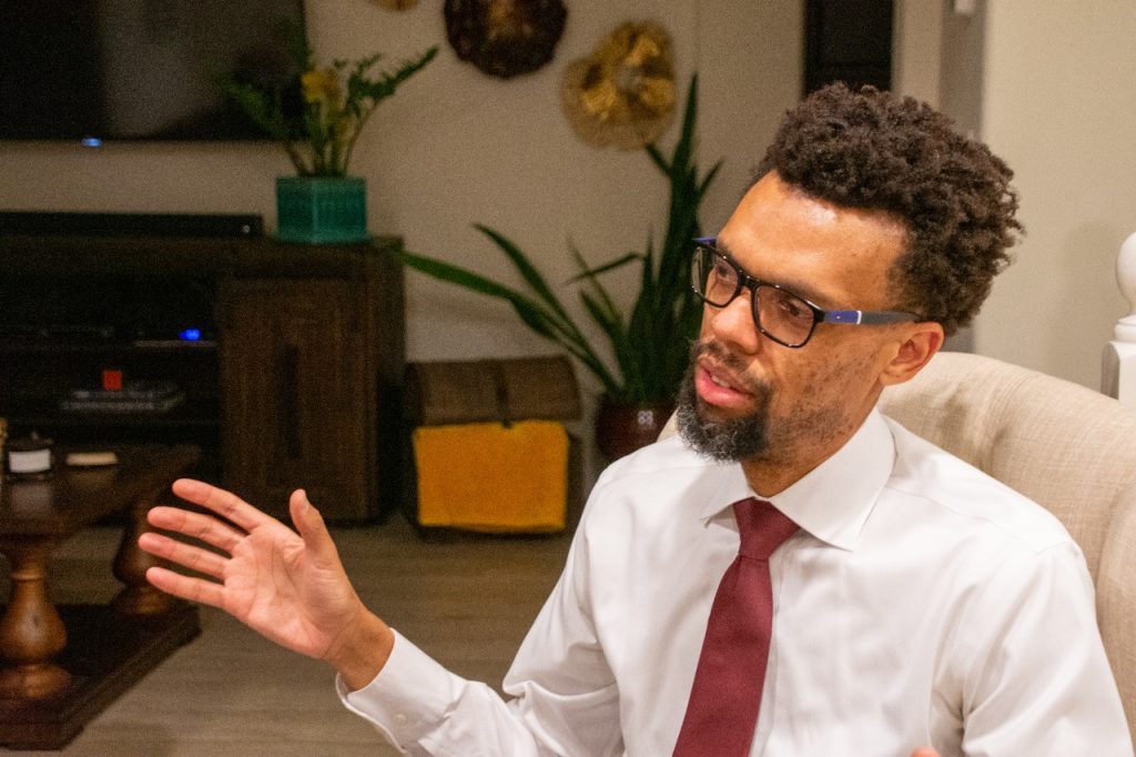 A man in glasses and white button-up shirt and red tie is talking with someone. His hand is up in a gesture.