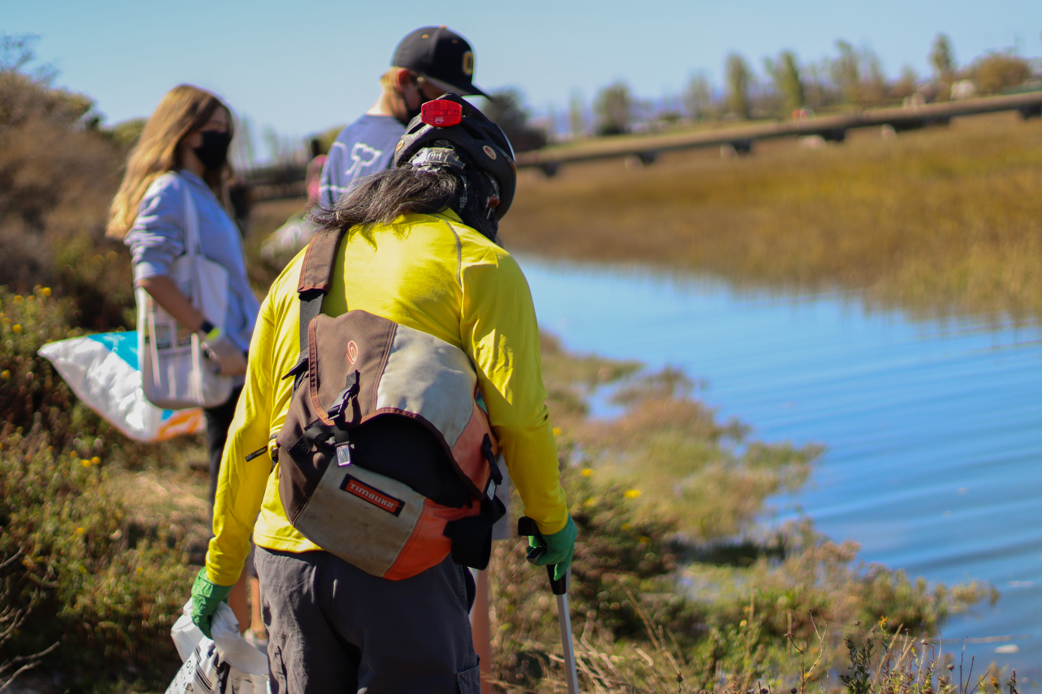 A woman is standing with two others picking up trash near a marsh.