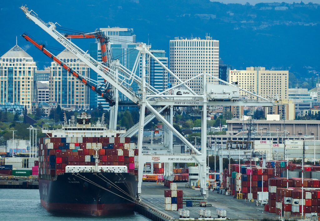 A container ship and cranes, with downtown Oakland and the Oakland Hills in the background.