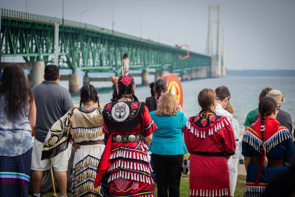 Indigenous ceremony being performed at the Straits of Mackinac. Photo of people's back in the foreground with the Mackinac Bridge in the background.
