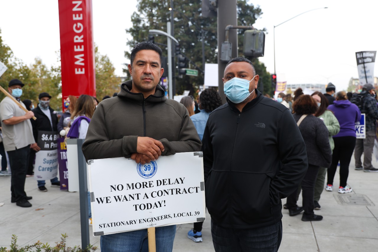 Two men stand side-by-side looking at the camera. Behind them is a crowd of people with signs.