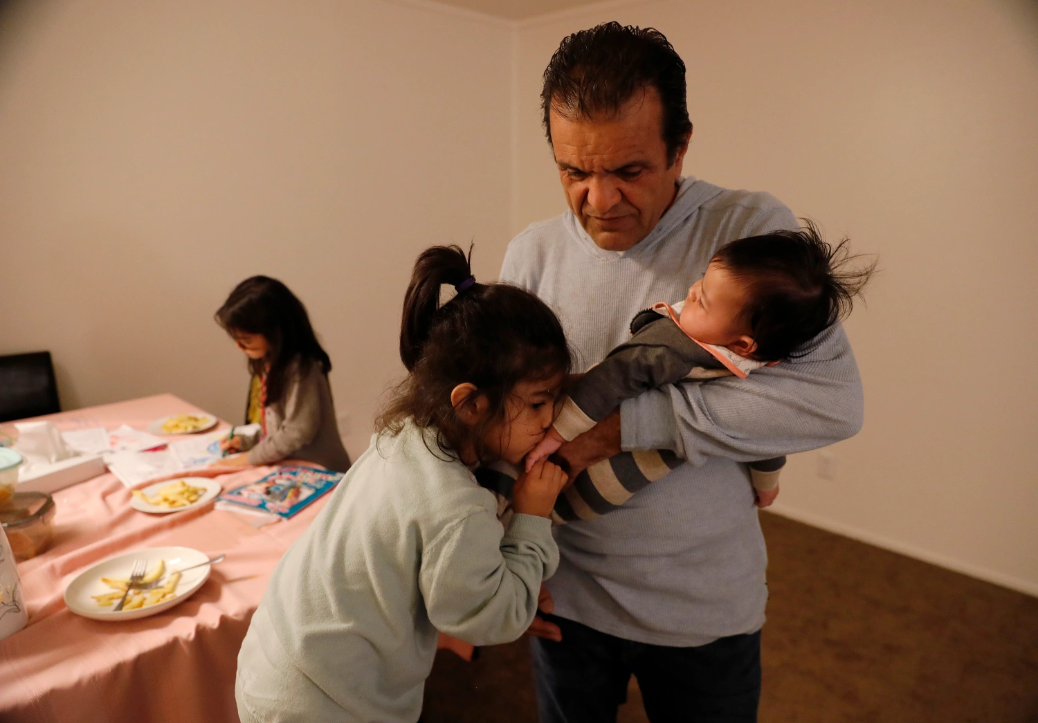A man is holding a baby, a young girl kisses the baby’s hand. Another girl is in the background at a table.