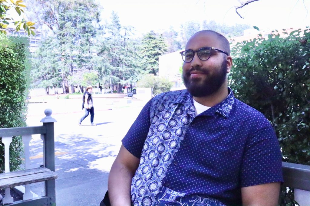 A man in a blue button shirt and glasses sits and smiles at the camera