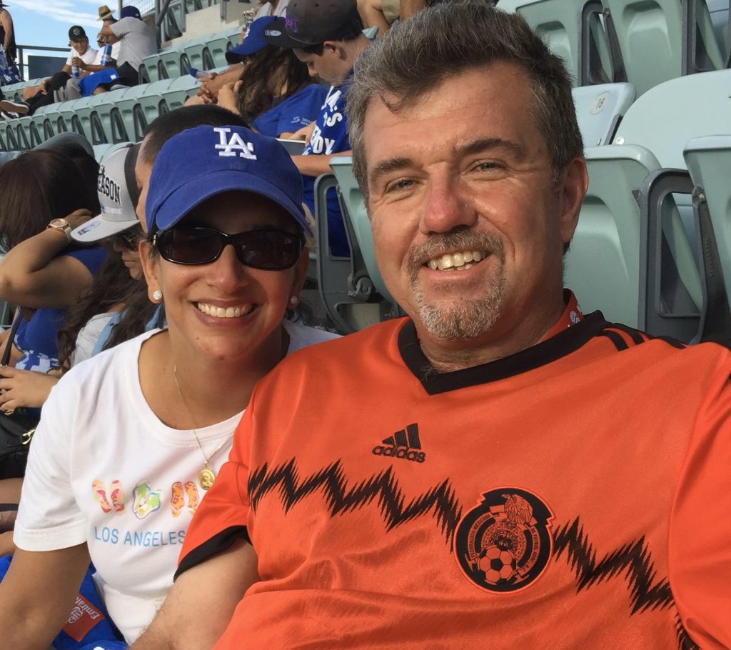 A woman wearing a blue LA Dodgers baseball and sunglasses smiles at the camera. A man wearing an orange shirt smiles at the camera next to the woman.
