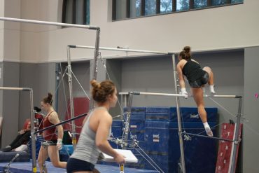 A picture of a gymnastics gym. There are uneven bars and several women who are doing various exercises.
