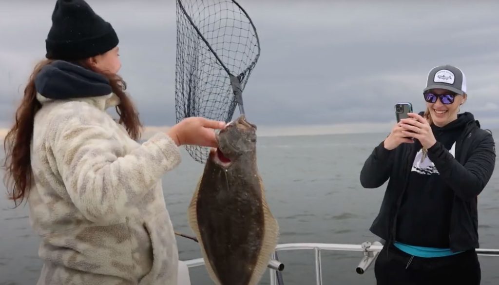 A woman with her back to the camera holds up a big fish. Another woman looking at the direction of the camera is taking a picture of the fisherwoman and her catch.