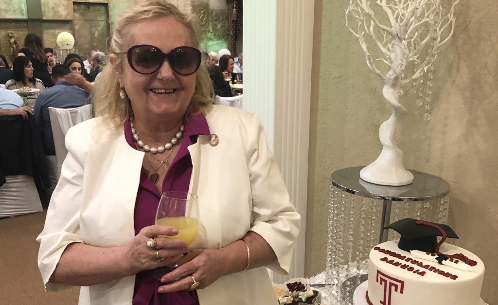 A woman in a dress and white blazer stands in front of a graduation cake.