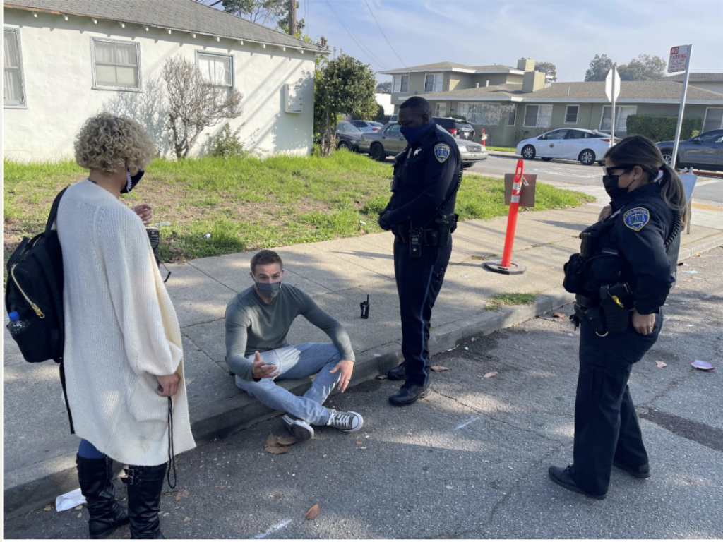 A man sits on a curb surrounded by two police officers and a clinician