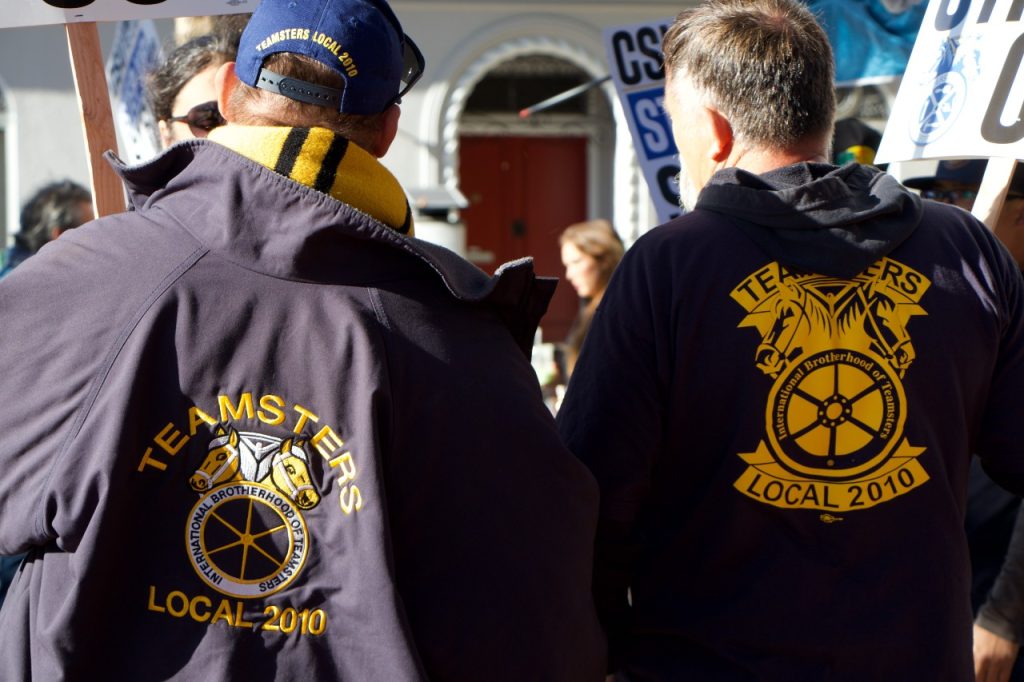 Two picketers stand side by side, thee teamsters logo showing on their jacket.