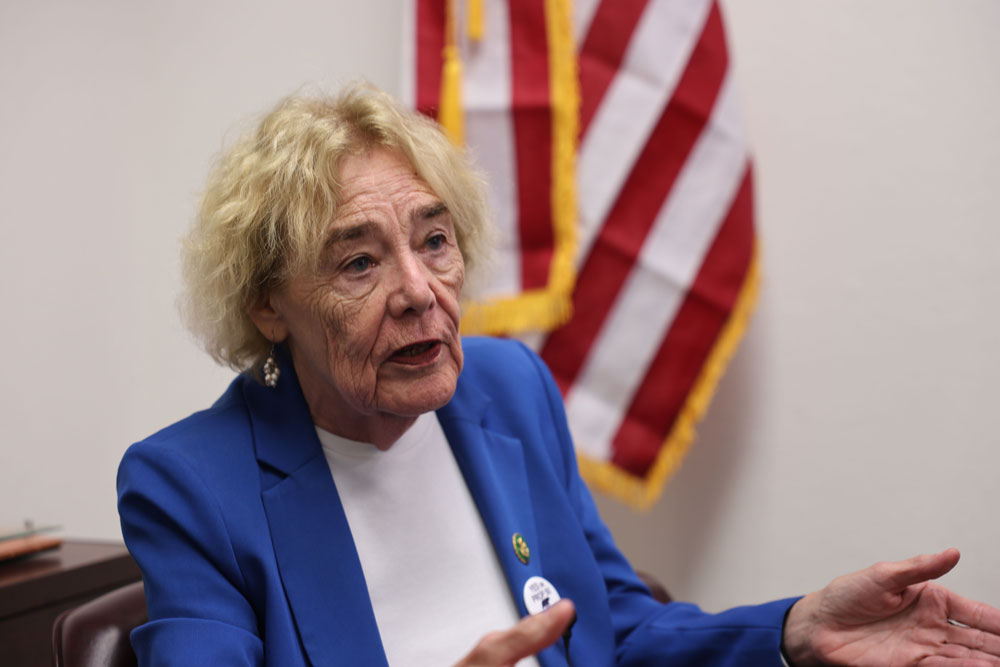 ET_2 Zoe Lofgren speaks during an interview, wearing a blue blazer and white shirt. She appears engaged in conversation, with an American flag partially visible behind her.