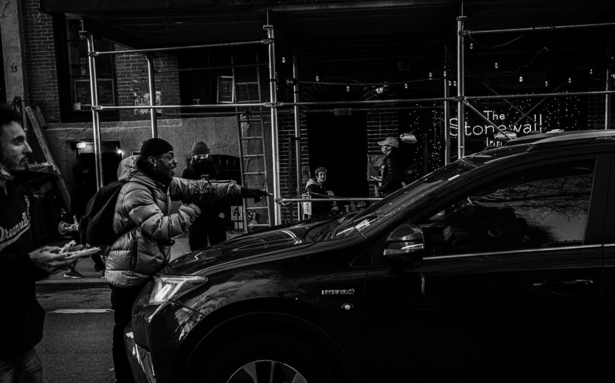 A protester is standing directly in front of a vehicle pointing at it. Other protesters stand by.